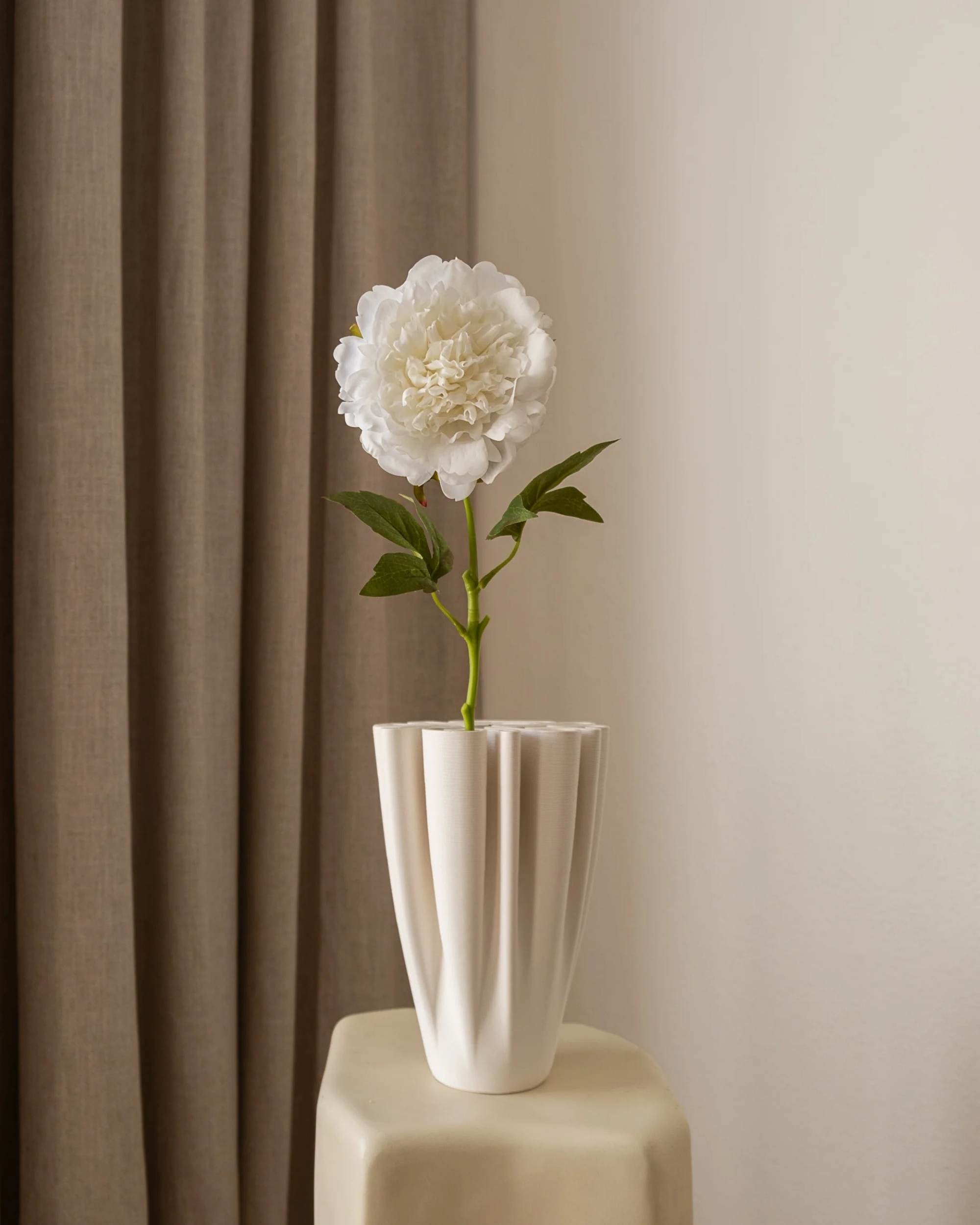 Amarante Peon i Hvit, kunstig blomst med grÞnne blader, i hvit plissert vase pÄ lyst bord mot gardin