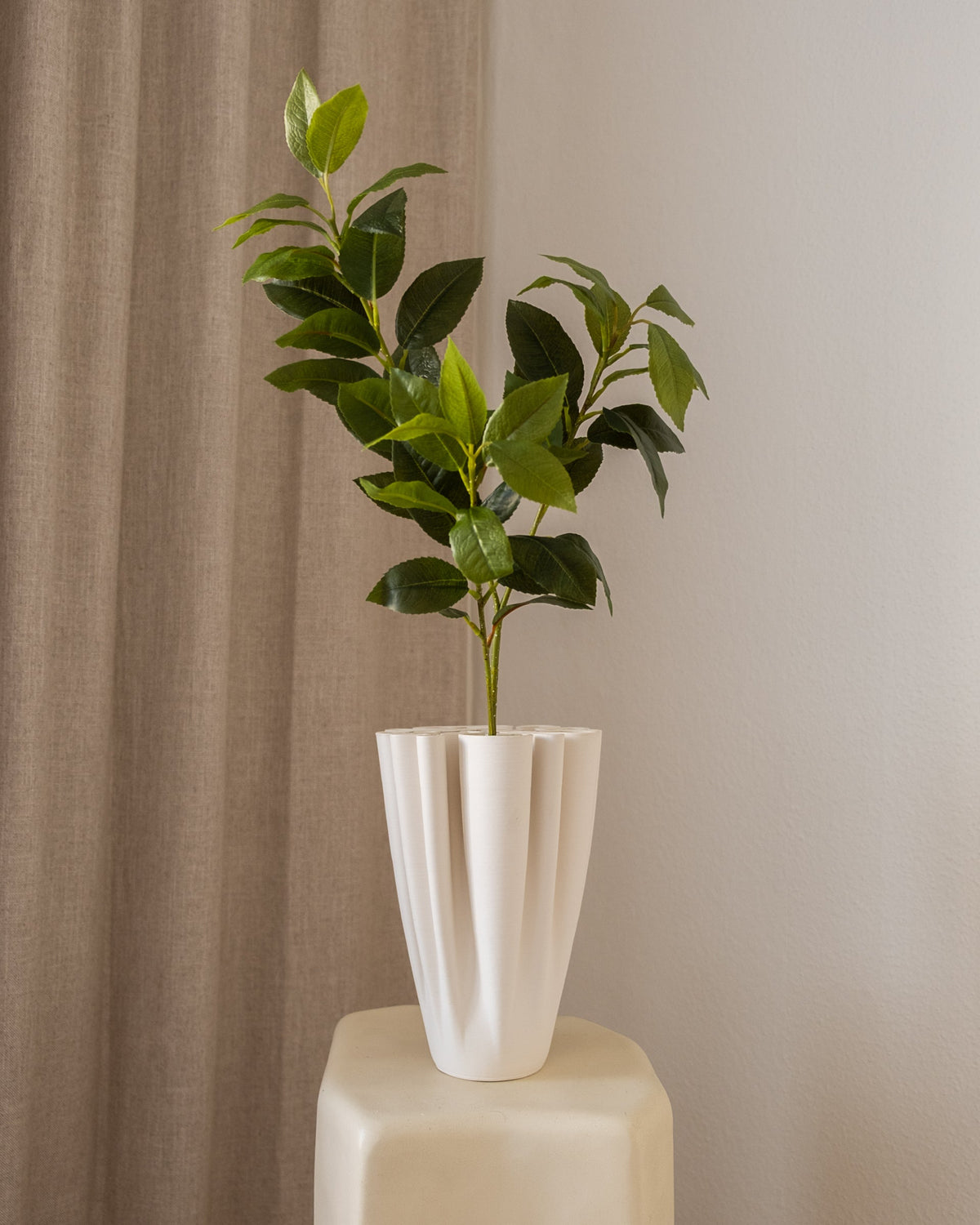 Amarante Osmanthus kunstig blomst med grønne blader i en hvit, plissert potte, mot en lys vegg