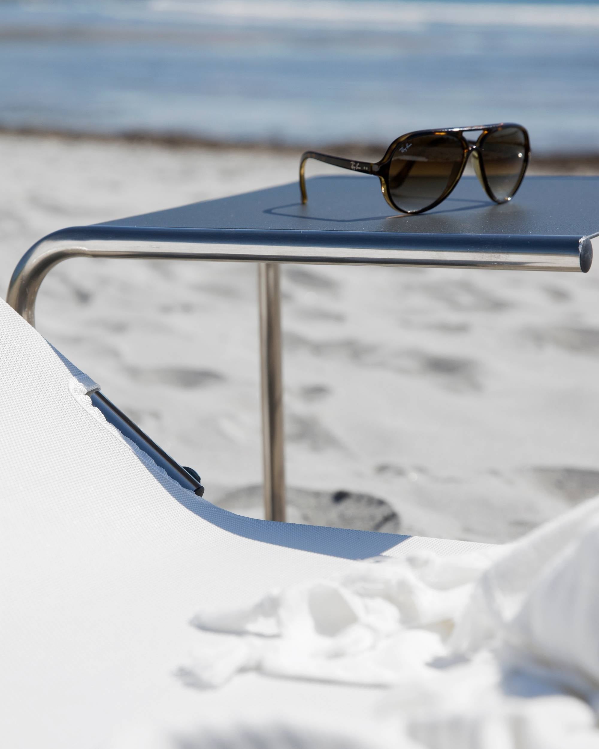 Fiam Club Table hagebord i antrasitt, med solbriller på, og en hvit strandstol i forgrunnen ved havet